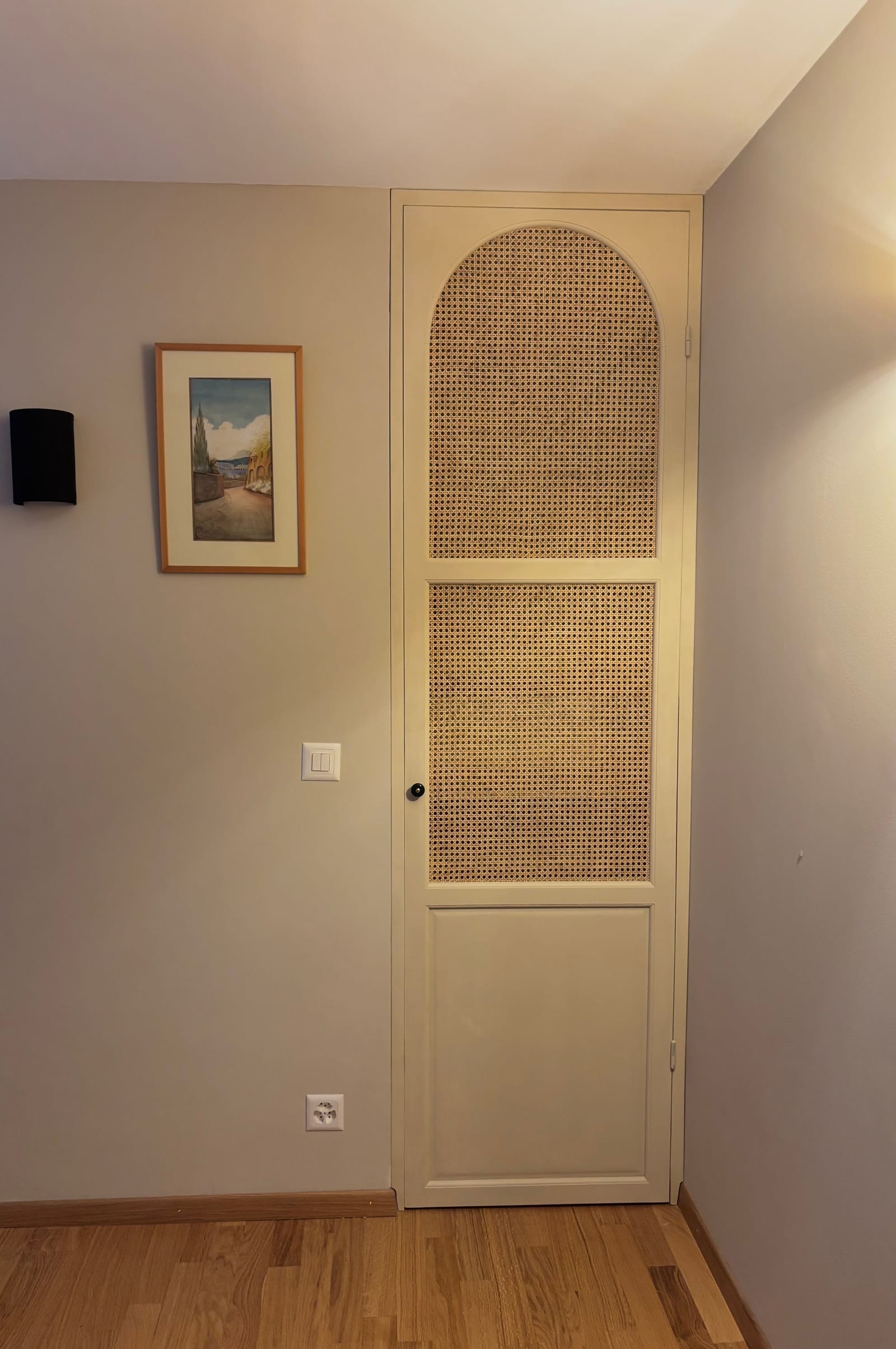 Porte en arche à deux panneaux de cannage et un panneau plein, posée dans un mur clair au-dessus d'un parquet.